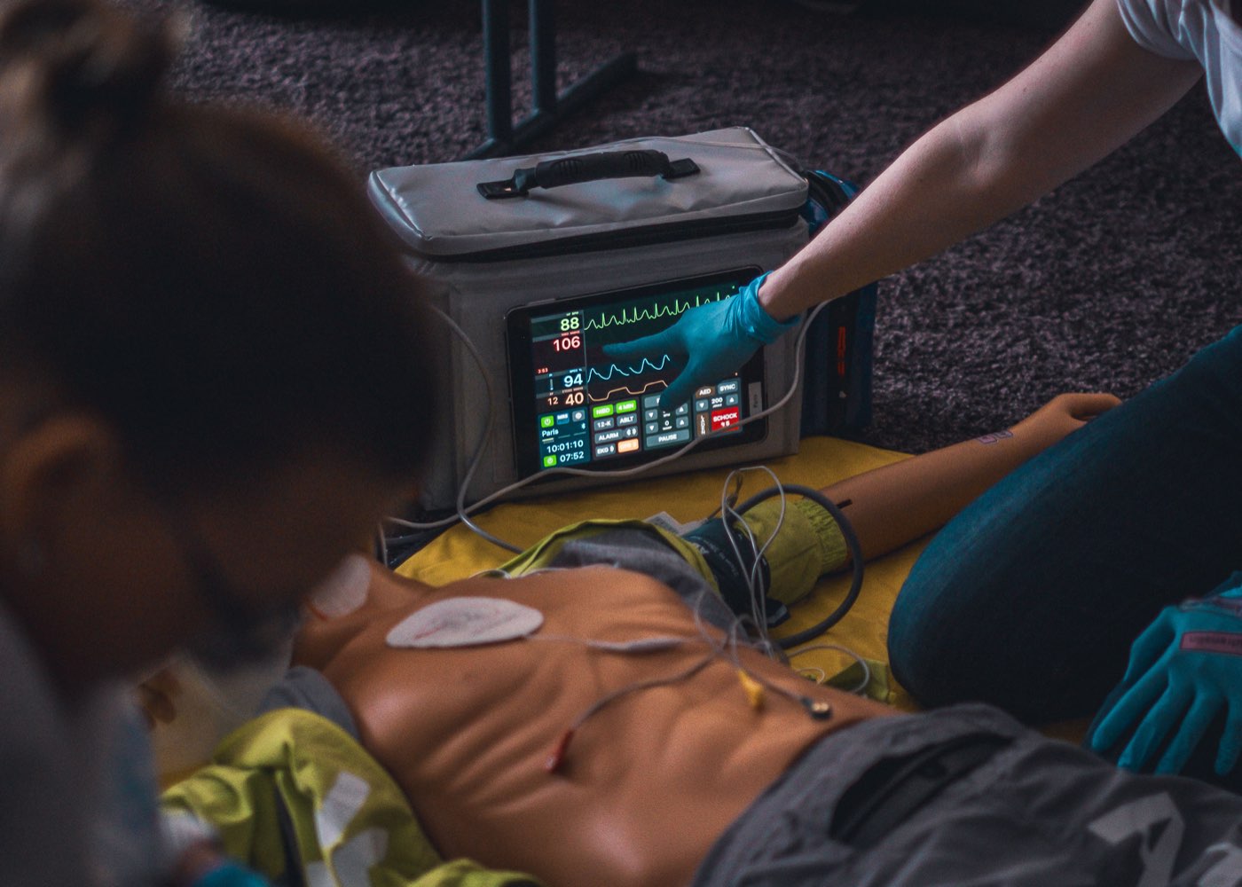 Eine behandschuhte Hand bedient den Monitor eines Defibrillators neben der Übungspuppe