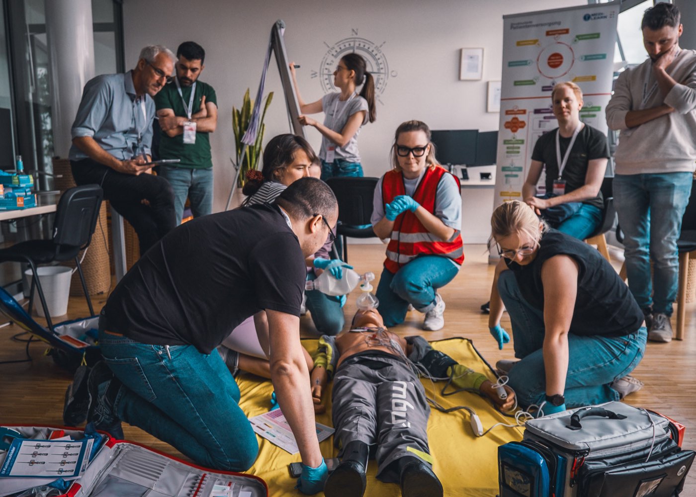 Megacode-Training: Ein Team arbeitet an der Übungspuppe, eine Instruktorin beobachtet vom Flipchart aus
