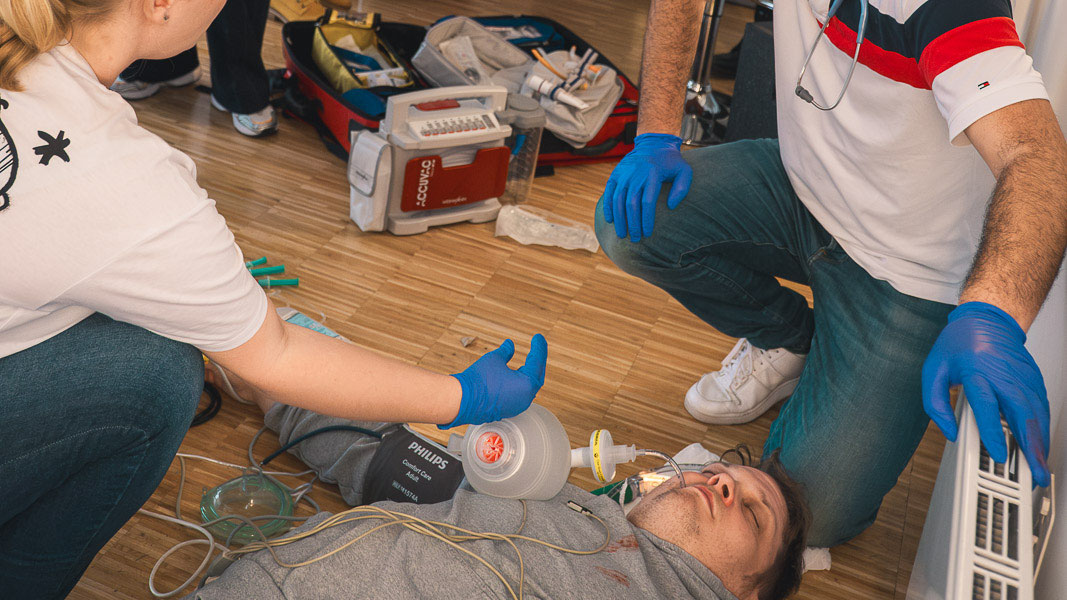 Zwei Teilnehmende knien bei einem am Boden liegenden Simulationspatienten, daneben Notfallrucksack und Beatmungsbeutel