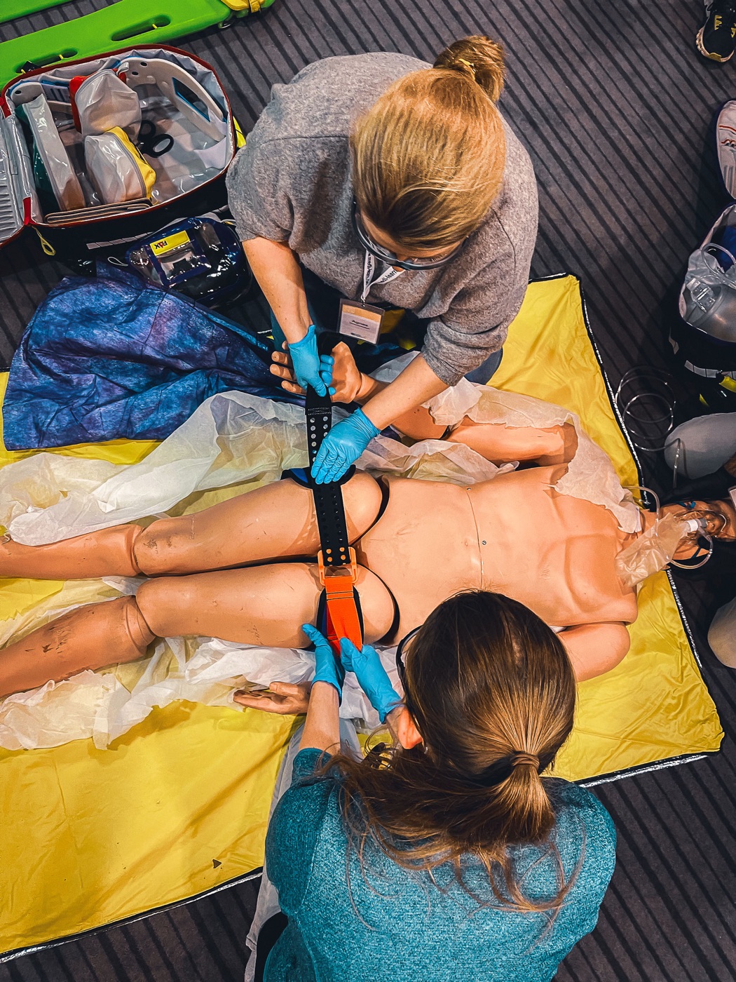 Zwei Teilnehmende legen einer Simulationspuppe eine Beckenschlinge an, Aufnahme von oben